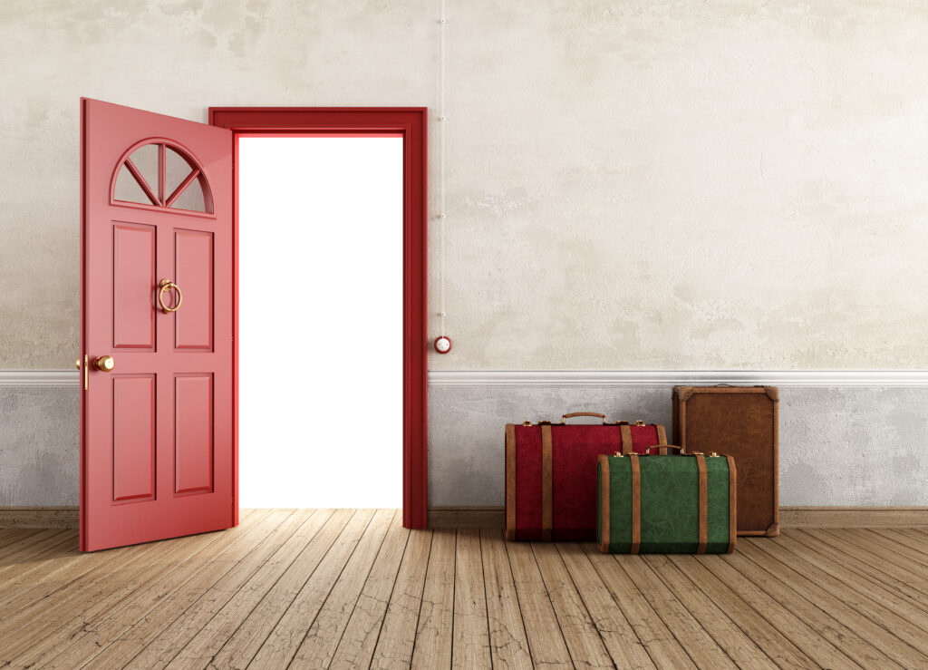 A trio of three vintage suitcases of different sizes and colors sits on a wooden floor to the right of an open red door.