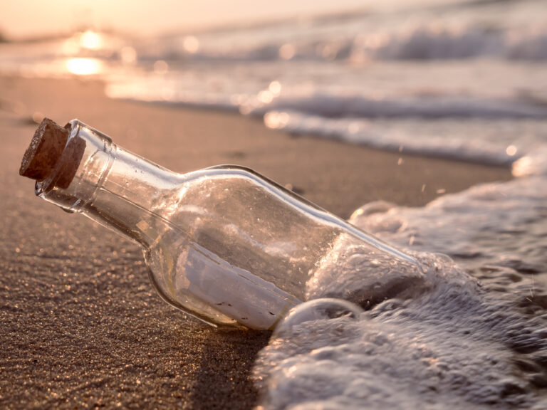 A clear glass bottle with a cork stopper lies on a beach in seafoam. In the bottle is a rolled-up note.